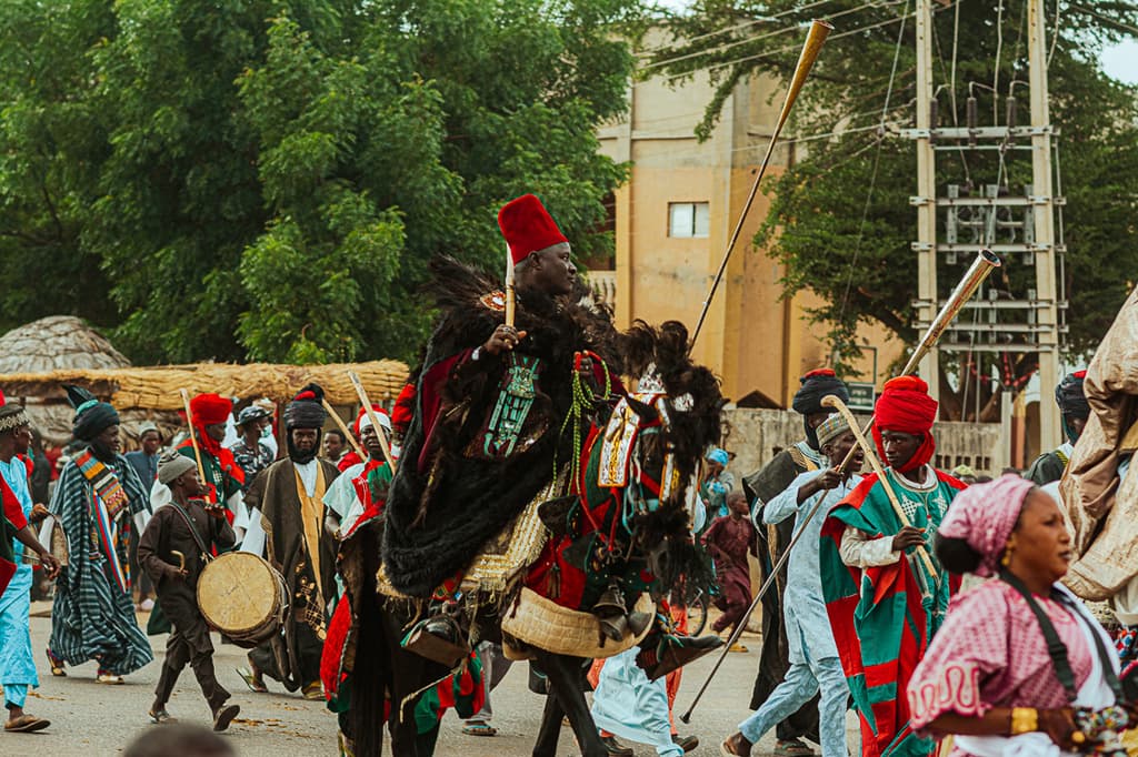 A turbaned Durbar horseman leads a ceremonial procession through the streets of Ilorin, surrounded by drummers and horn players.