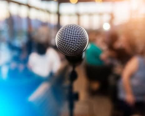 A stage microphone in sharp focus with a warmly lit audience blurred behind it.